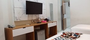 Habitación de hotel con un escritorio de madera, un televisor montado encima, un refrigerador pequeño, una cama con un edredón estampado y un espejo.