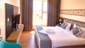 A hotel room with a bed, a desk, a chair, a ceiling fan, and a view through the window. - Antilia Inn (Badami)