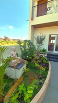 Una estatua de una figura sentada en un jardín con diversas plantas y un edificio con balcón al fondo.