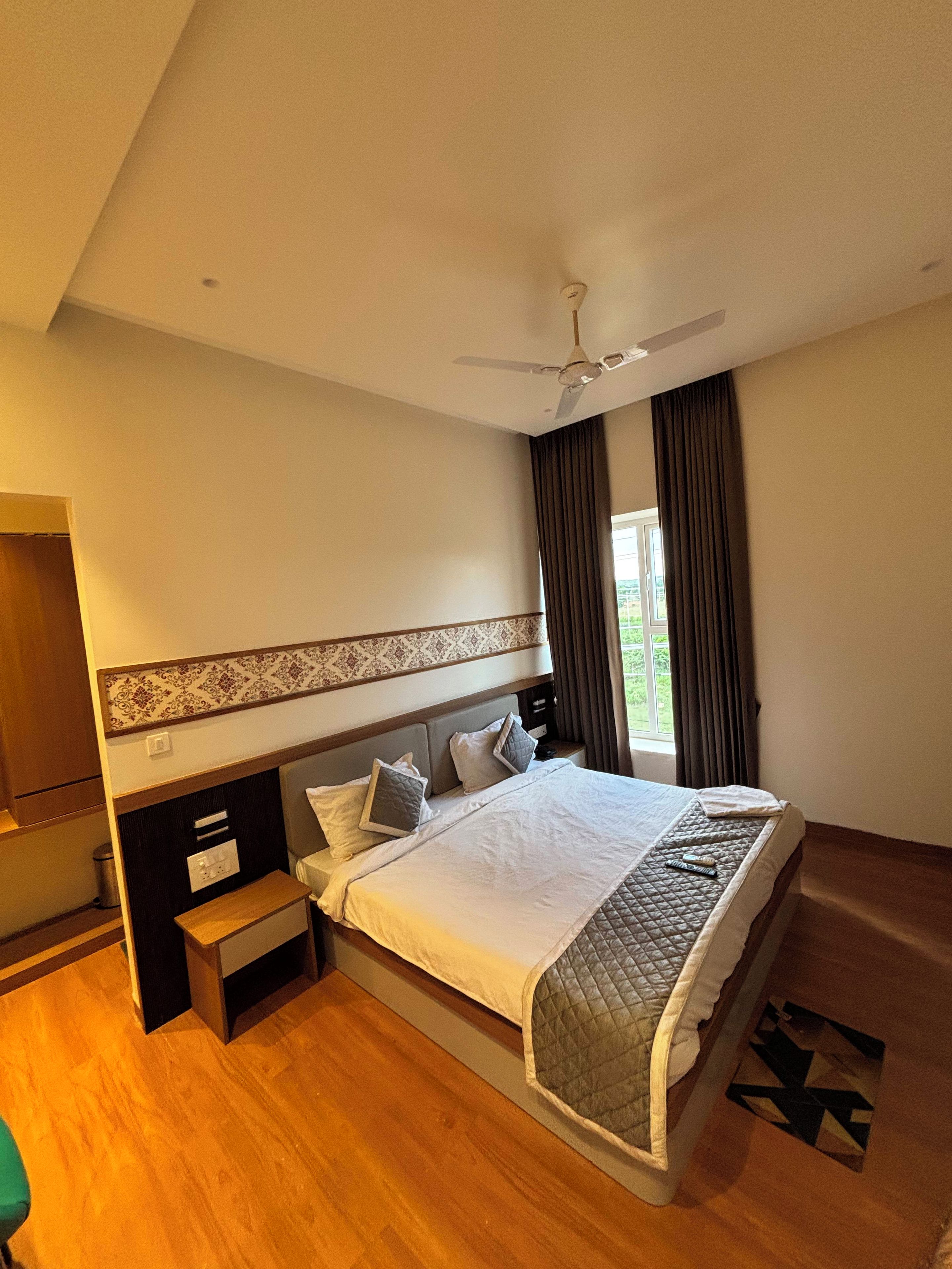 A hotel room with a large bed, a ceiling fan, a window with curtains, a wooden floor, and a small bedside table.