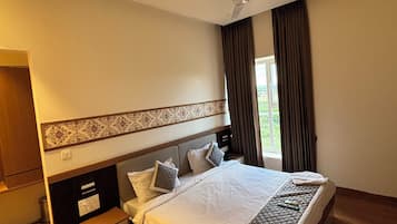 A hotel room with a large bed, a ceiling fan, a window with curtains, a wooden floor, and a small bedside table.