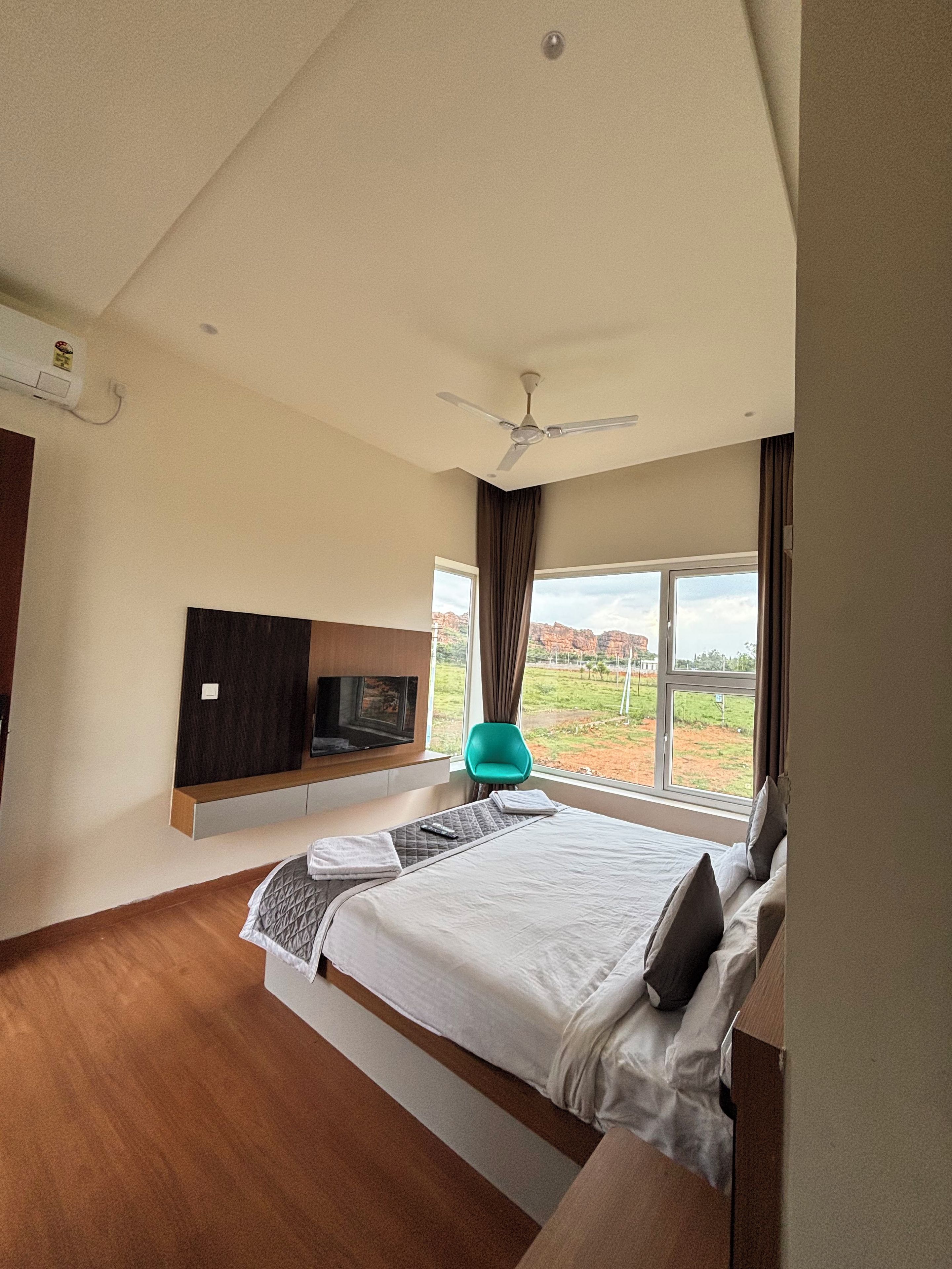 A modern hotel room with a large bed, a flat-screen TV mounted on the wall, a ceiling fan, and a view of the outdoors.