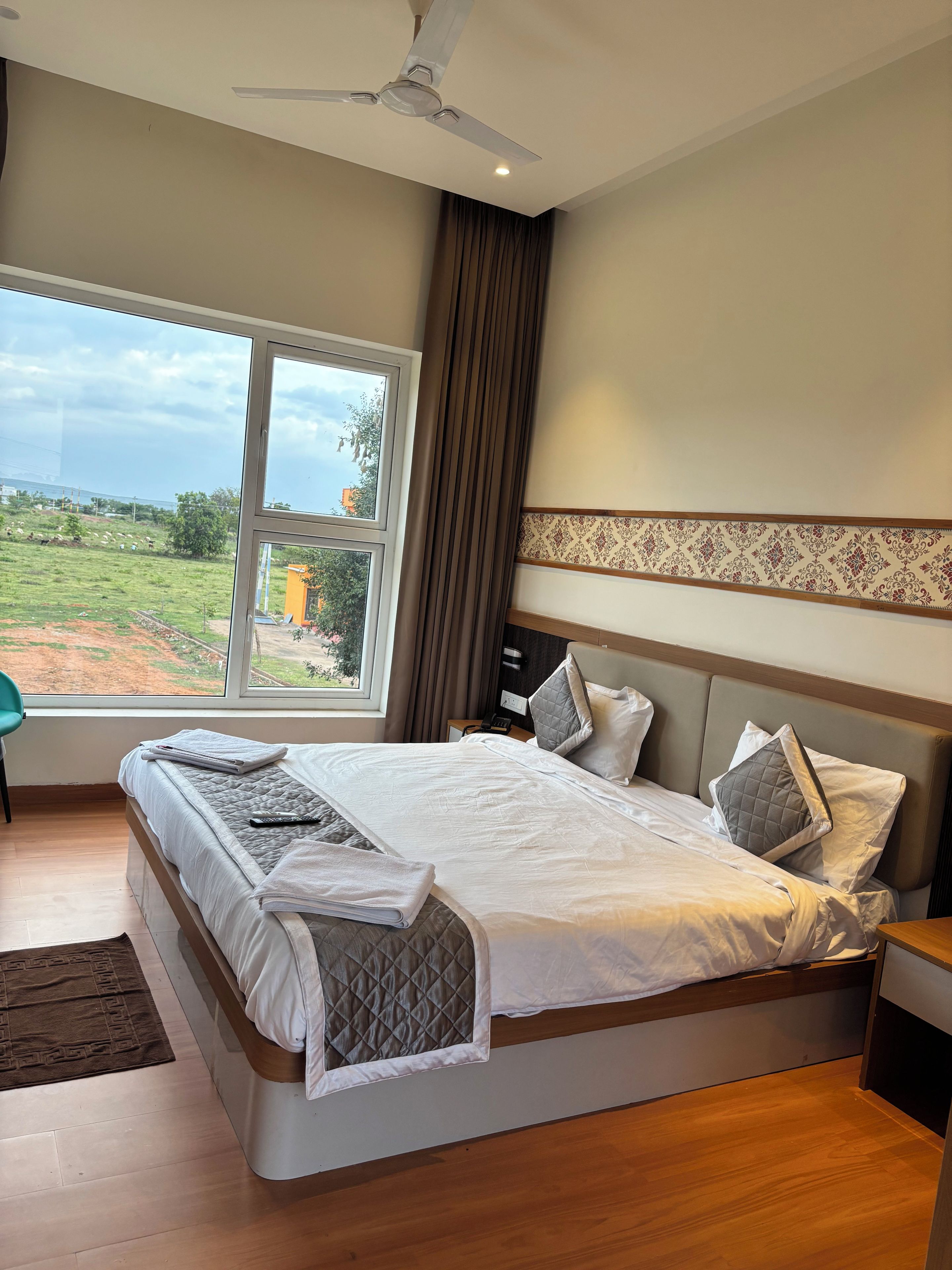 A hotel room with a large bed, a ceiling fan, a view of the outdoors, and a bedside table with a lamp.