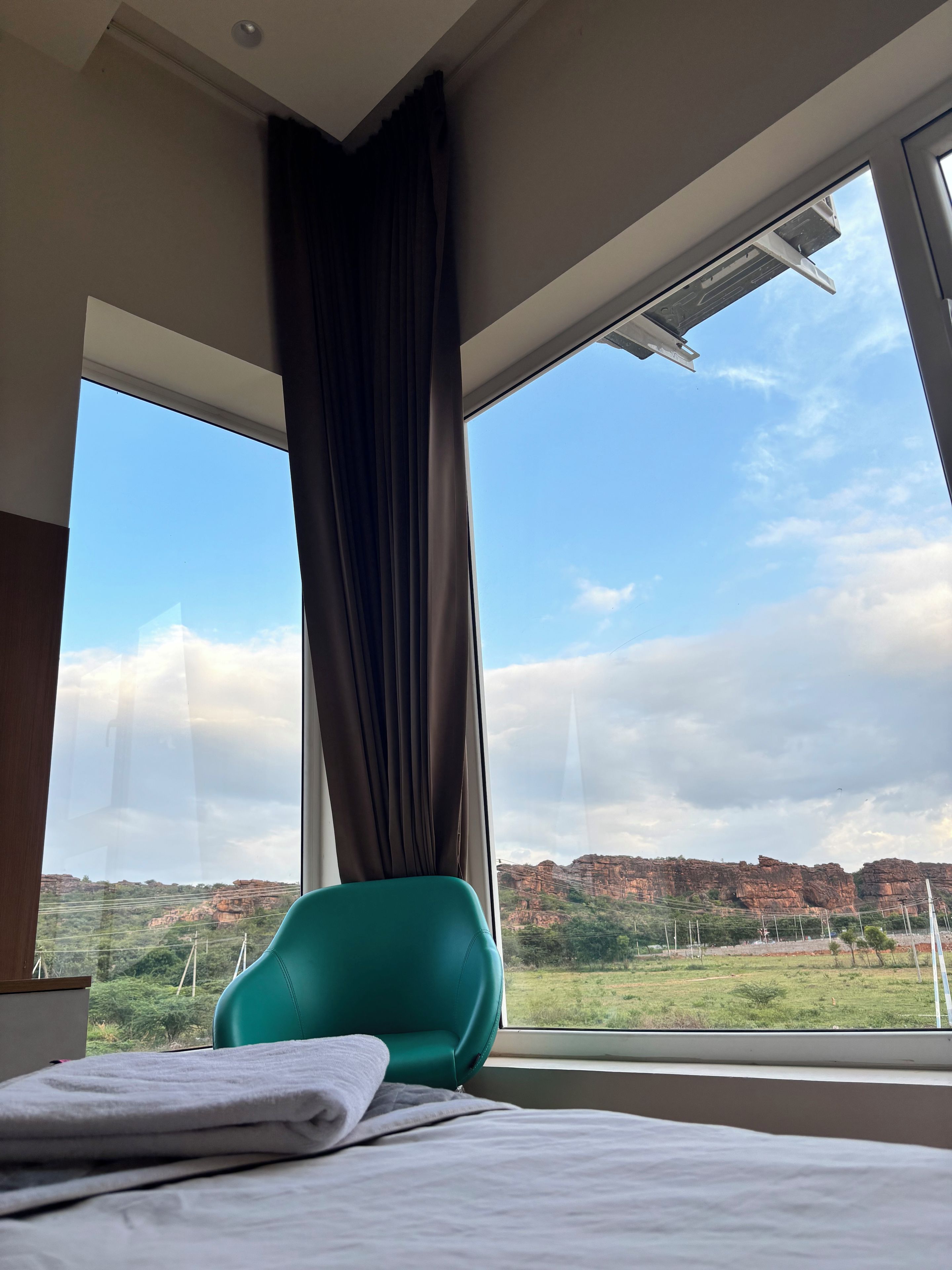 A hotel room with a bed, a teal chair, and a large window offering a view of a mountainous landscape.