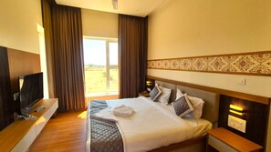 Quarto de hotel com uma cama, televisão, mesa de cabeceira de madeira e janela com cortinas.