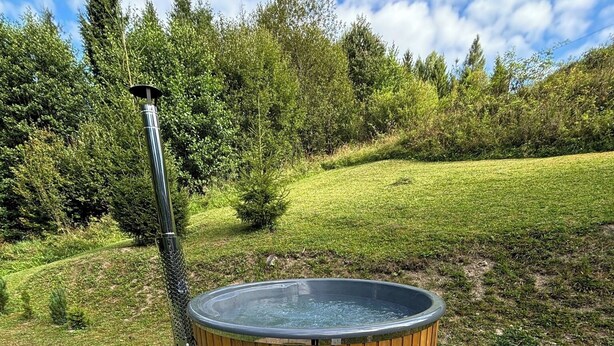 A hot tub with wooden steps and a metal pole, surrounded by a stone patio, set in a grassy area with trees in the background.
