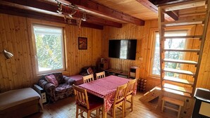 A wooden cabin interior with a dining table, chairs, a sofa, a TV, and a ladder.