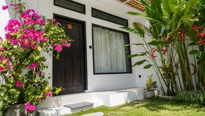 A modern building with a dark door, a window with a curtain, and a garden with pink flowers and green plants. - Susurro Villas (Kuta)