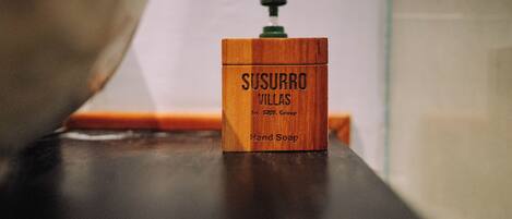 Un dispensador de jabón de madera con la etiqueta 'SUSURRO VILLAS by S.A.P.O. Group' sobre una superficie oscura.