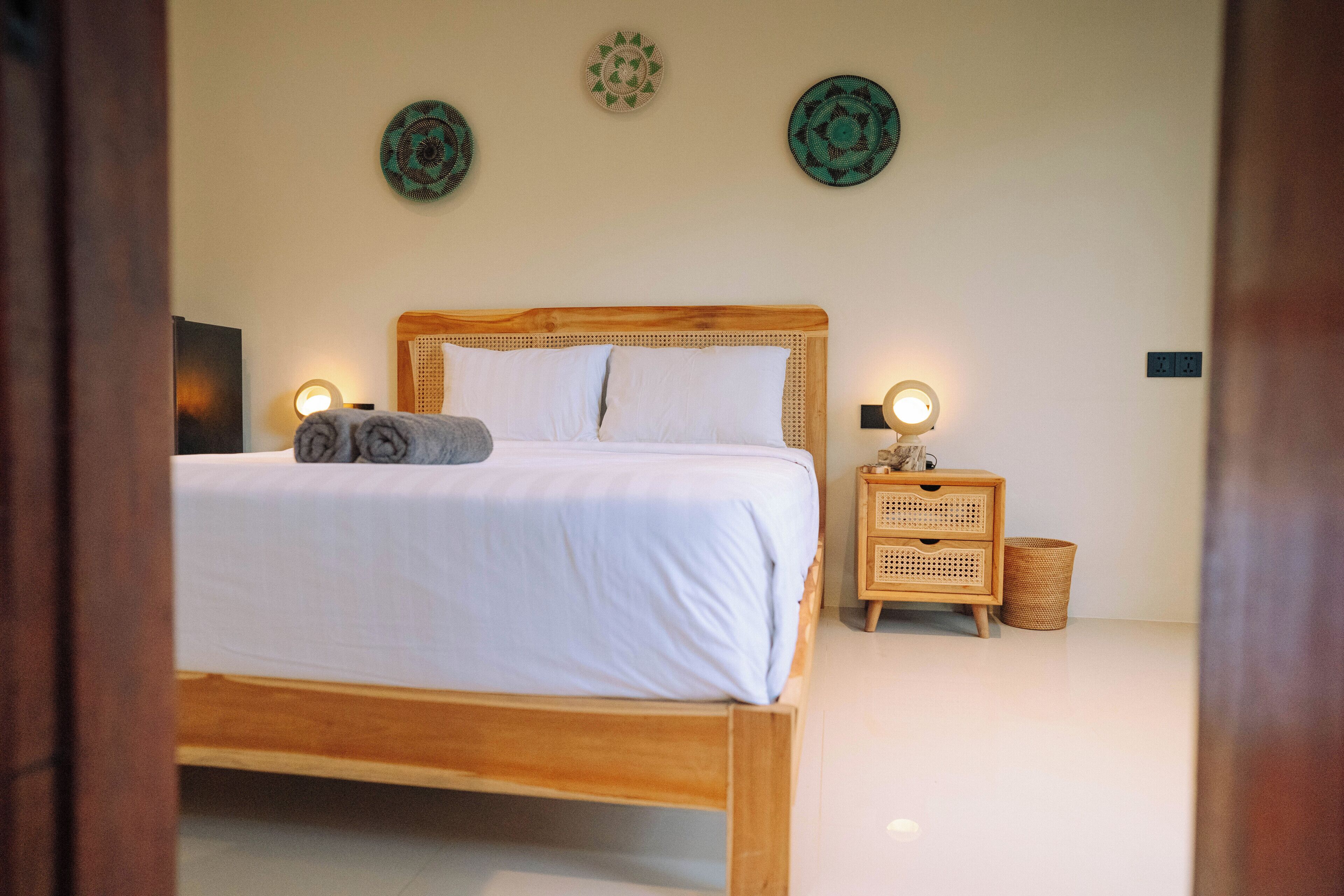 A neatly made bed with white linens and two pillows, a wooden headboard, a bedside table with a lamp, and a woven basket.