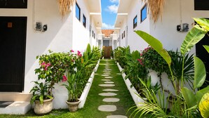 A narrow, white corridor with a central green path, flanked by potted plants and thatched-roof structures. - Susurro Villas (Kuta)