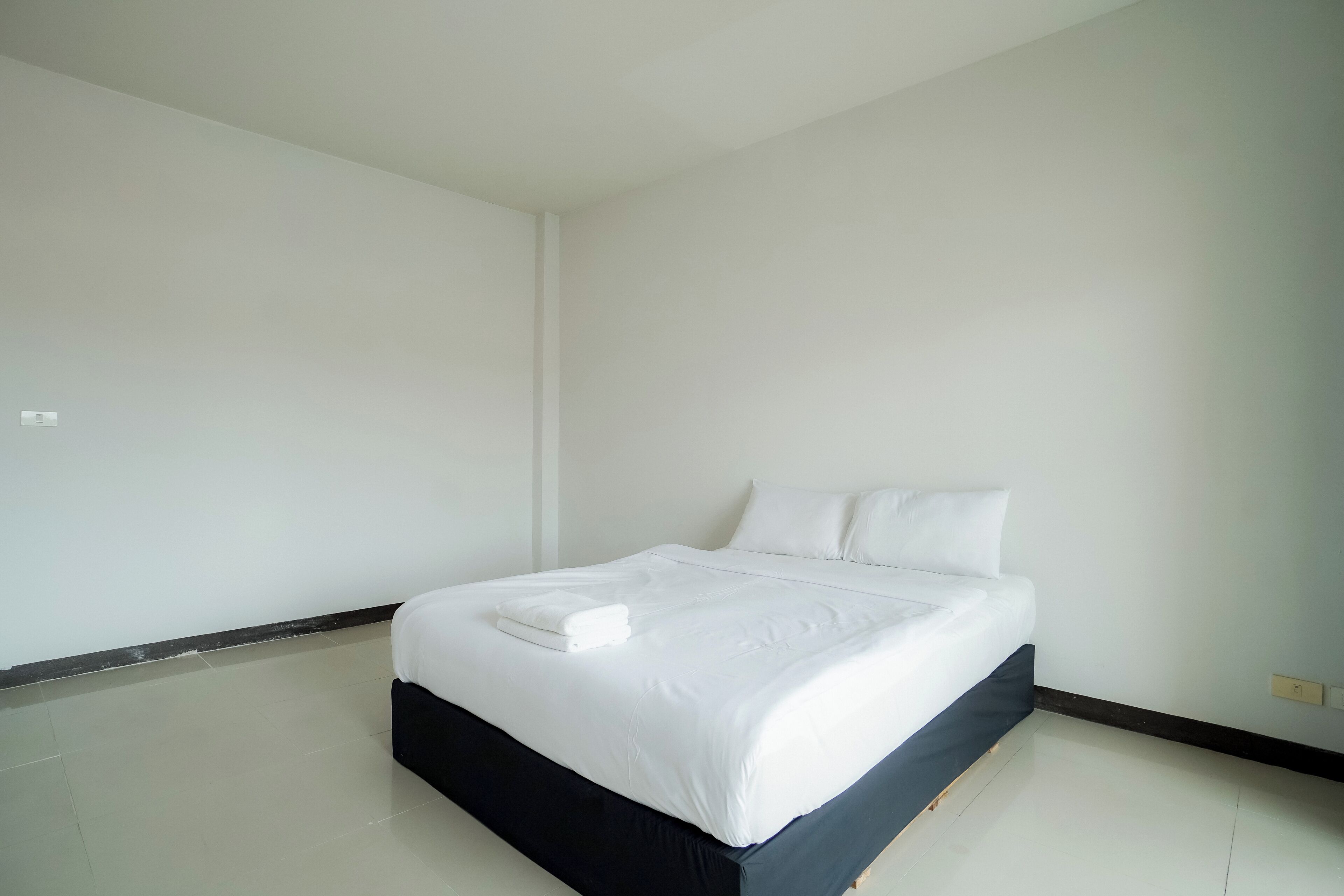 A clean, minimalist bedroom with a large bed, white bedding, and a dark-colored headboard.