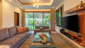 A modern living room with a sofa, coffee table, TV, and large windows. - ELIVAAS Azalea 3 BHK with Pvt Pool (Siolim)