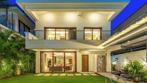 A modern two-story house with a balcony, large sliding glass doors, and a well-maintained lawn. - ELIVAAS Azalea 3 BHK with Pvt Pool (Siolim)
