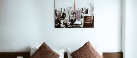 Habitación de hotel con cama, dos almohadas marrones, una colcha blanca, una obra de arte con paisaje urbano en la pared y un cabecero de madera.