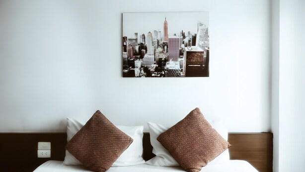 A hotel room with a bed, two brown pillows, a white comforter, a cityscape artwork on the wall, and a wooden headboard. - Snap Inn Thaphae (Chiang Mai)
