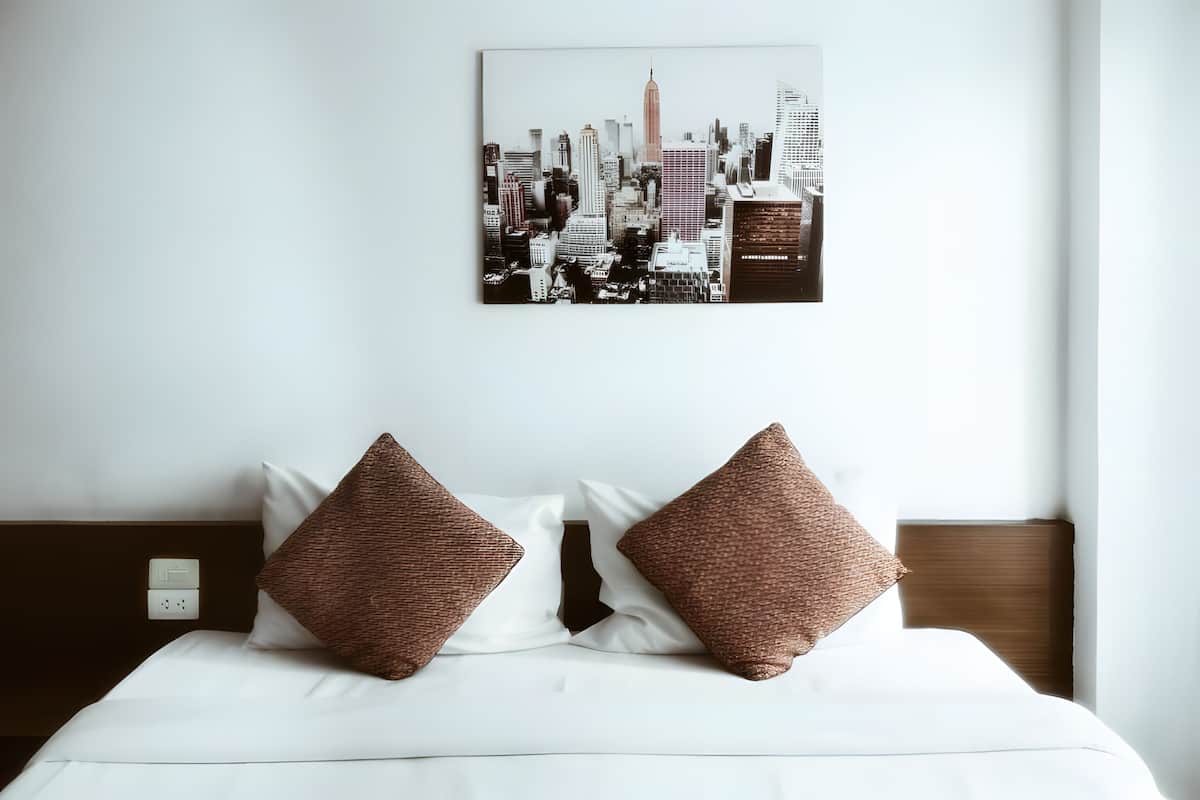 Ett hotellrum med en säng, två bruna kuddar, ett vitt täcke, ett konstverk med en stadsbild på väggen och en sänggavel i trä.