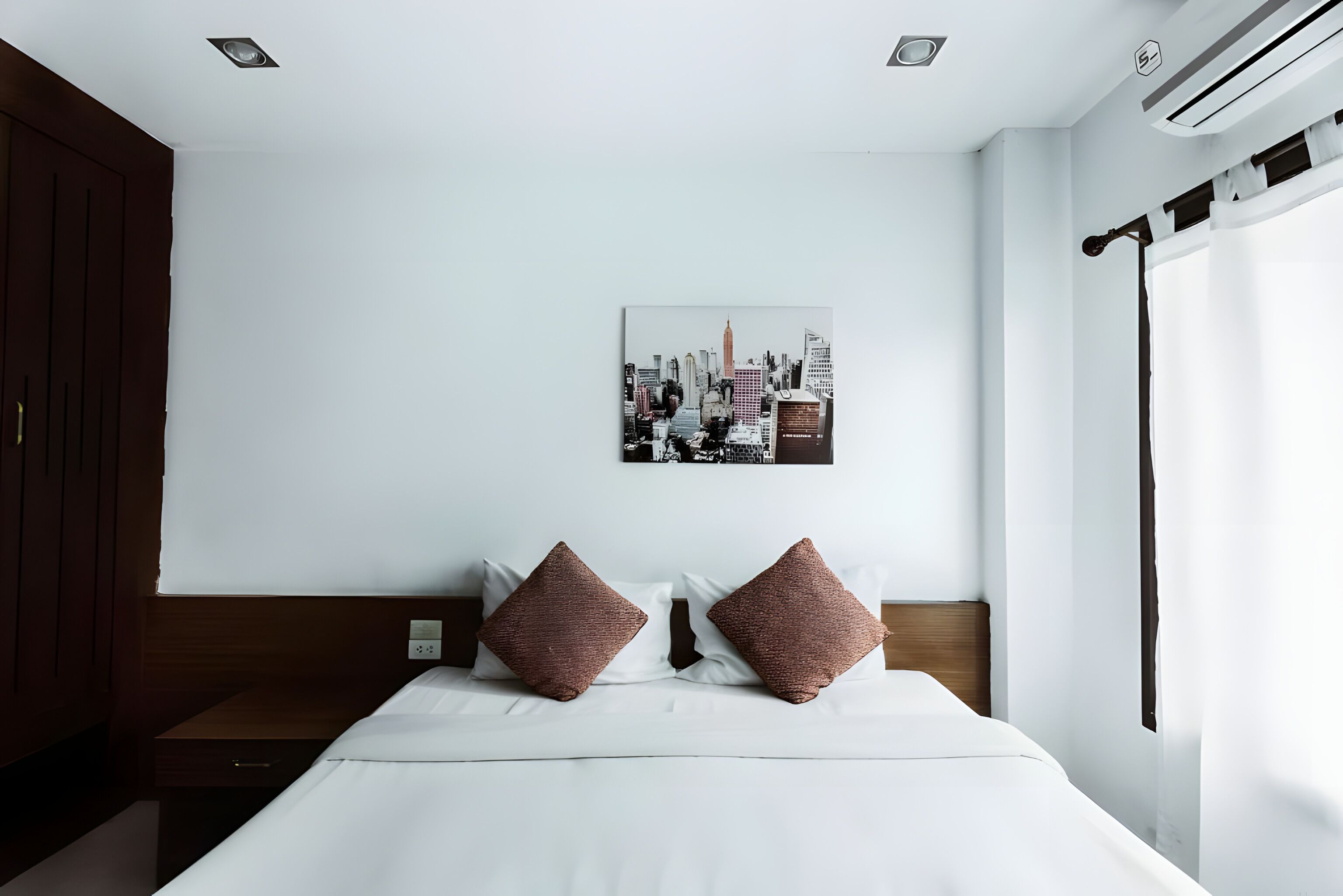 Une chambre d’hôtel avec un lit, une œuvre d’art représentant un paysage urbain accrochée au mur, une tête de lit en bois et une fenêtre avec des rideaux.