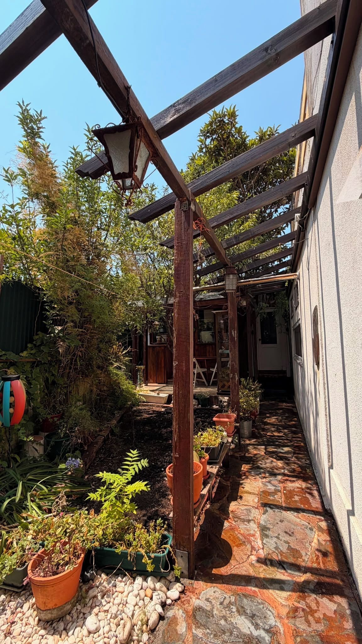 Uma estreita trilha de pedra leva a uma pérgula de madeira com um lampião, rodeada por plantas em vasos e um jardim.