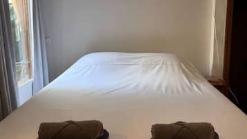 Quarto de hotel com uma cama grande, duas toalhas, uma cruz na parede e uma janela com cortinas.