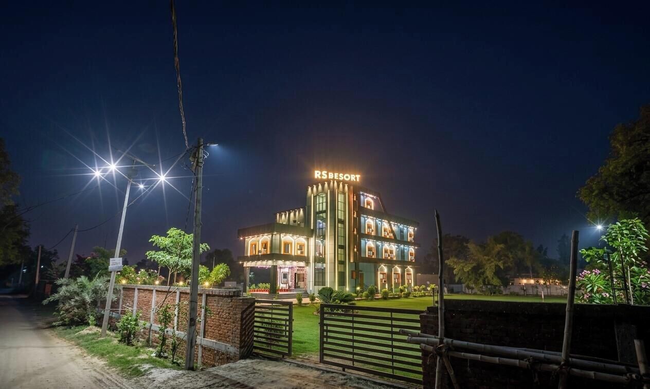 Un immeuble à plusieurs étages, sur lequel on peut lire l’inscription « RSRESORT » en haut, entouré d’un jardin bien éclairé et doté d’une entrée fermée par un portail.