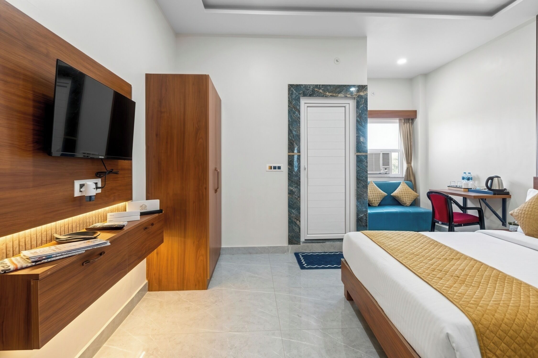 Een hotelkamer met een bed, een tv bevestigd aan een houten paneel, een bureau met een stoel, en een deur die naar een andere kamer leidt.