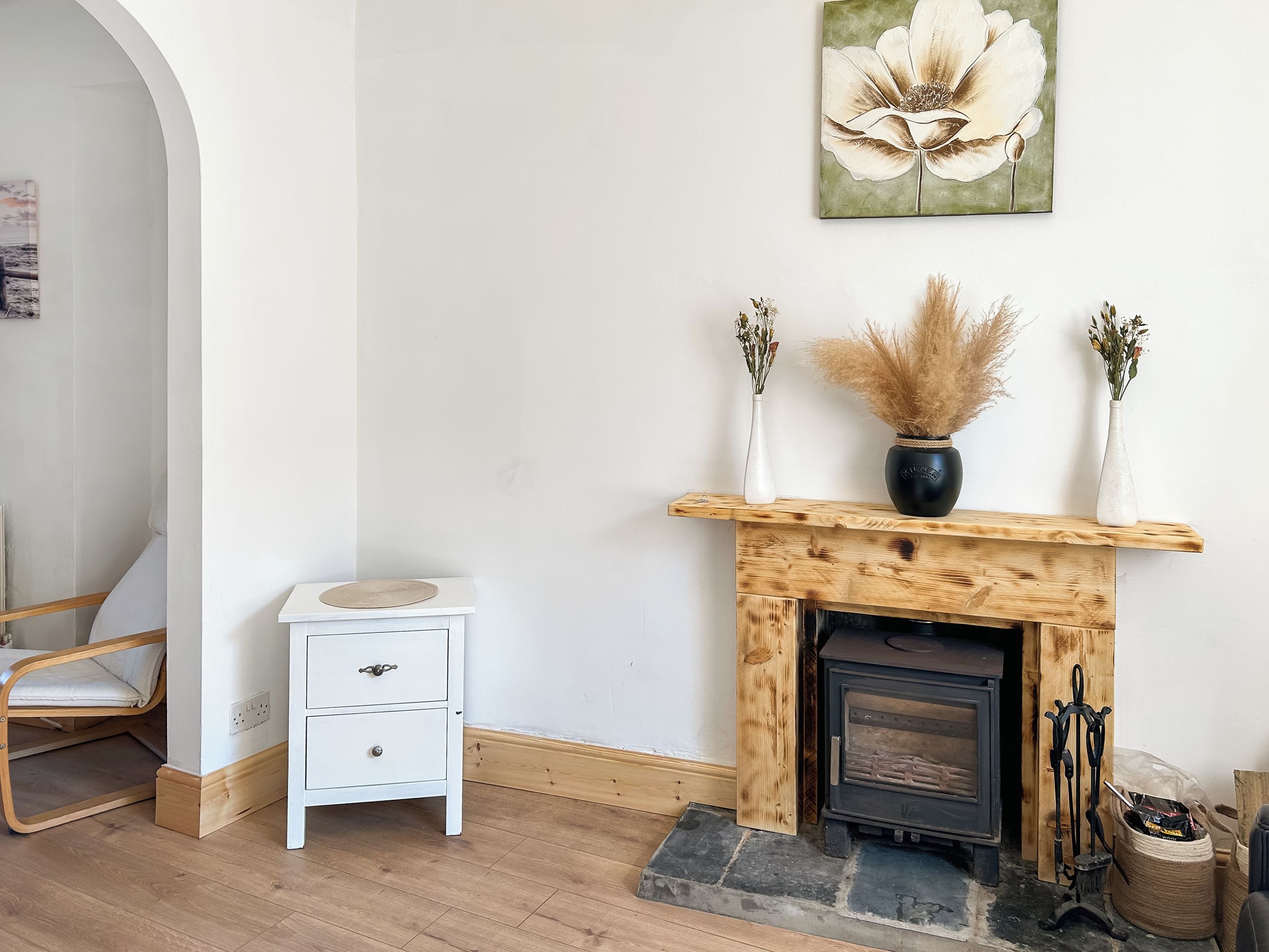 A room with a white dresser, a wooden mantelpiece with a vase and dried plants, a fireplace, and a large floral painting on the wall.
