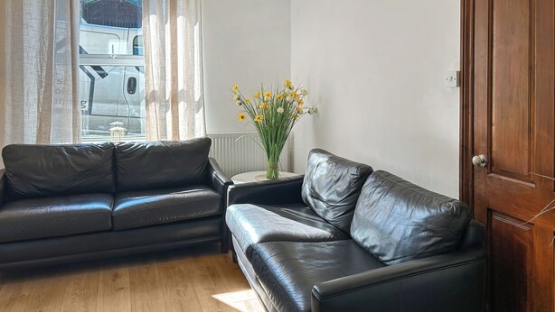Un salon avec un canapé en cuir noir, une petite table sur laquelle est posé un vase de fleurs et un plancher en bois.