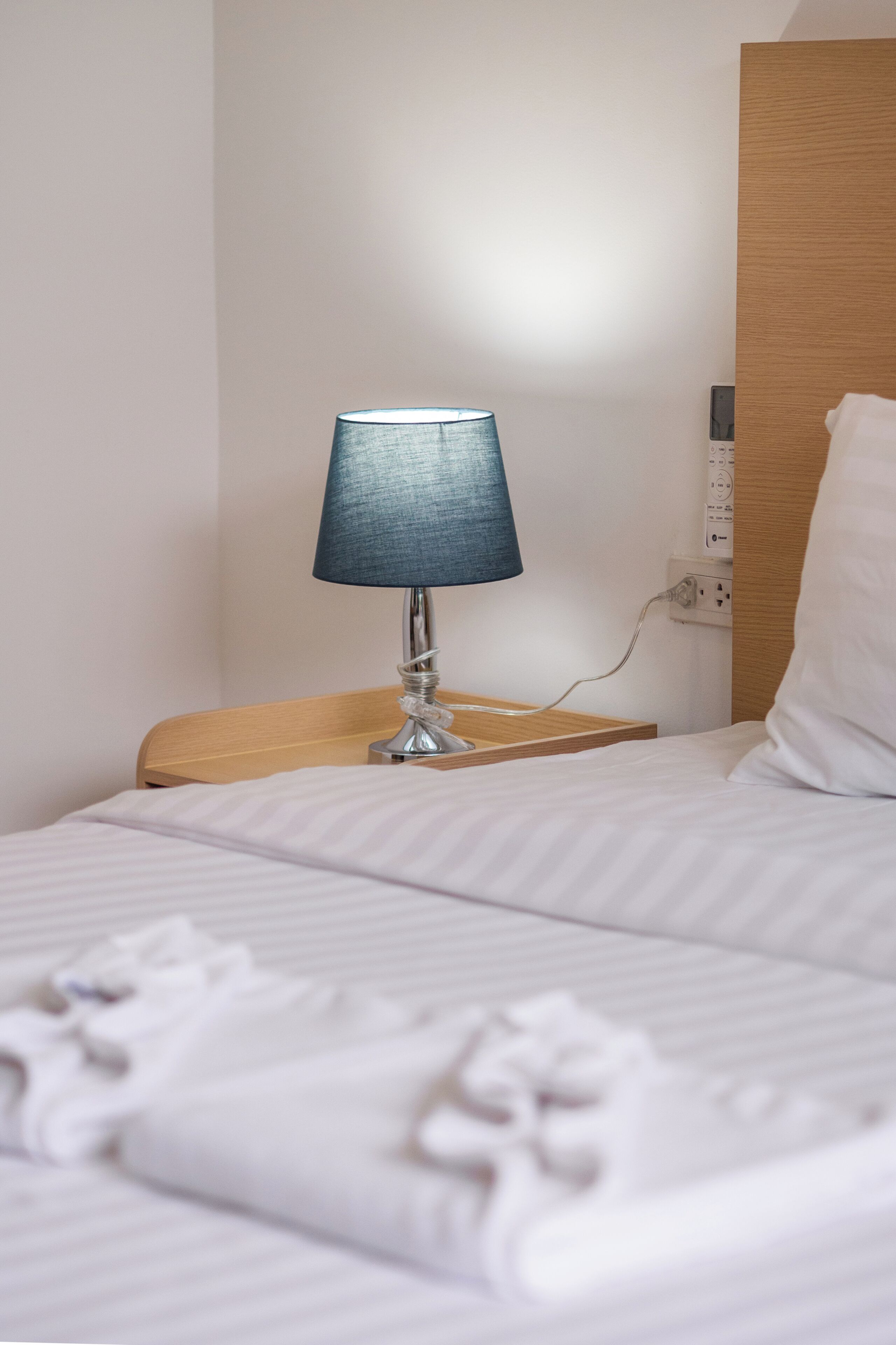 En pænt redt seng med hvid dyne og puder, et sengebord med en lampe, og en stikkontakt på væggen.