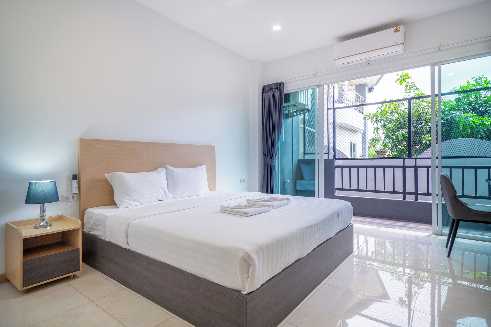 A modern hotel room with a large bed, a bedside table with a lamp, a balcony with a chair, and a view of greenery outside.