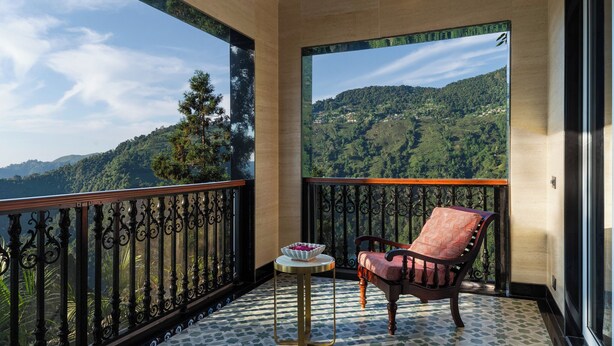 Un balcon donnant sur un paysage montagneux, une petite table ronde avec un saladier, et une chaise rouge avec un coussin.