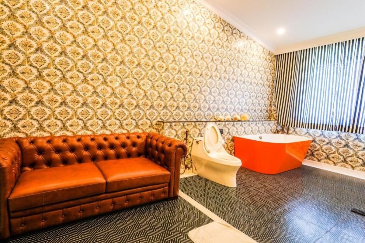 Une salle de bain avec un papier peint à motifs, un canapé marron capitonné, des toilettes blanches et une baignoire rouge.