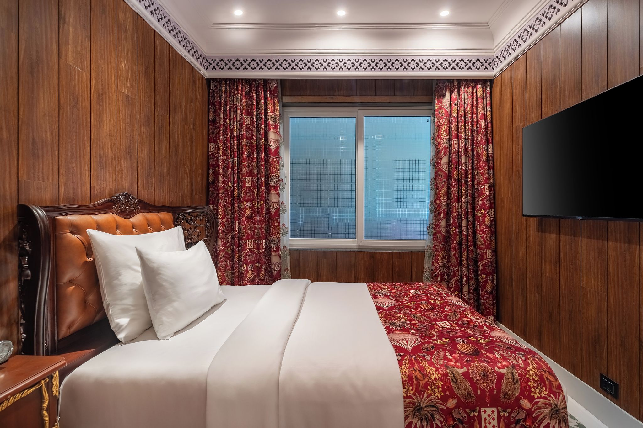A hotel room with a wooden headboard, a large bed with white bedding, red patterned curtains, and a TV on the wall.