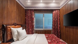 Een hotelkamer met een houten hoofdeinde, een groot bed met witte beddengoed, rood met patroon bedekte gordijnen en een televisie aan de muur.