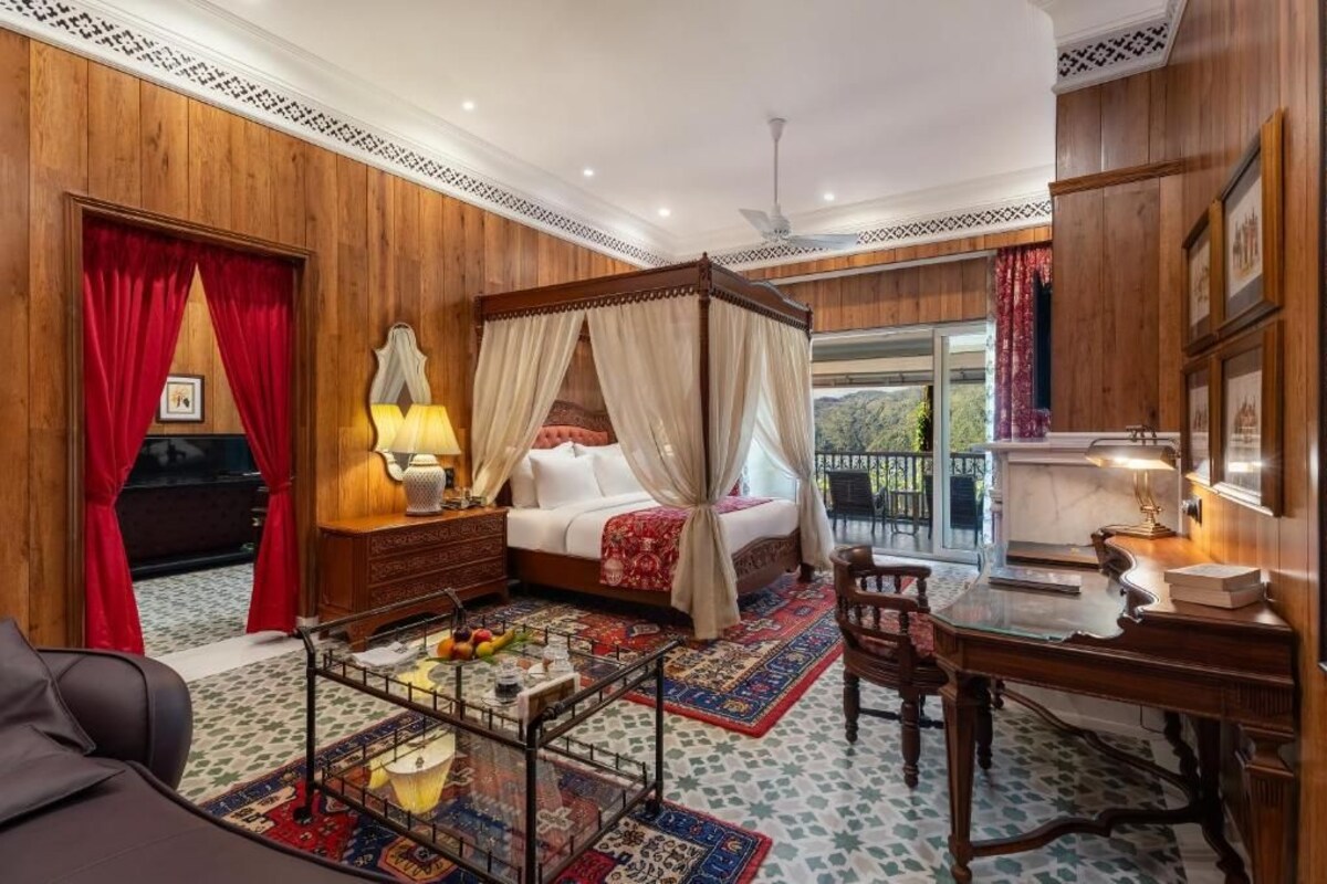 Quarto com cama de dossel, escrivaninha, cadeira, mesinha de cabeceira com abajur e vista pela janela.