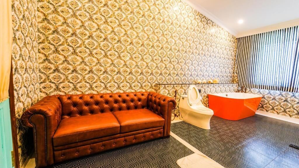 Une salle de bain avec un papier peint à motifs, un canapé marron capitonné, des toilettes blanches et une baignoire rouge.