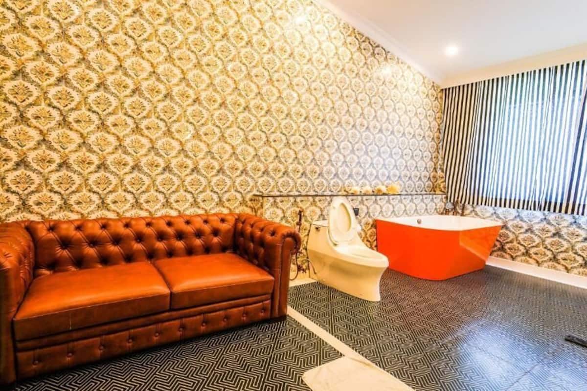 Une salle de bain avec un papier peint à motifs, un canapé marron capitonné, des toilettes blanches et une baignoire rouge.