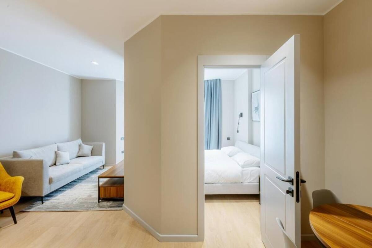 Een moderne hotelkamer met een bank, een houten bijzettafeltje, een bed met witte lakens en een deur naar een andere kamer.