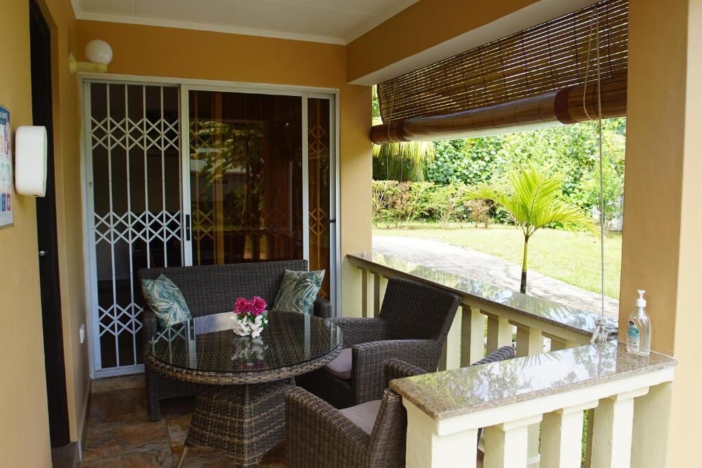 A covered outdoor seating area with wicker furniture, a glass table, and a view of a garden.