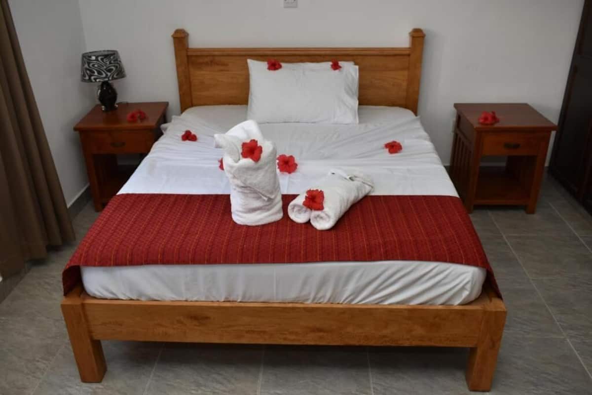 Un lit bien fait, recouvert d’une couverture rouge, avec des oreillers blancs et des fleurs rouges. Deux tables de chevet en bois, chacune surmontée d’une lampe et d’un petit objet décoratif, se trouvent de part et d’autre du lit.