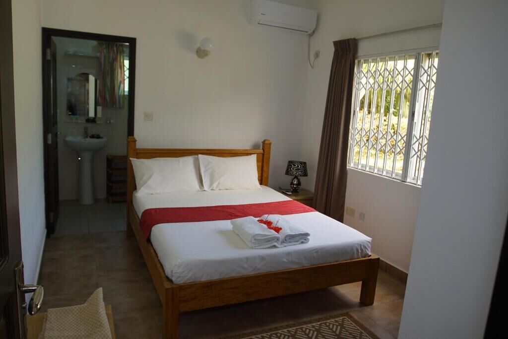 A bedroom with a wooden bed, a bedside table, a mirror, a window with curtains, and an air conditioning unit.