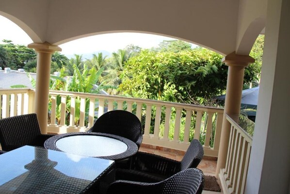 A covered outdoor seating area with a table and chairs, a white railing, and lush greenery in the background. - Zeph Self Catering Apartment (Mahé Island)