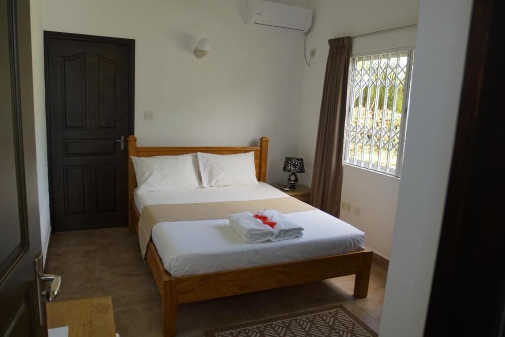 A hotel room with a bed, a door, an air conditioning unit, a nightstand, and a window with curtains.