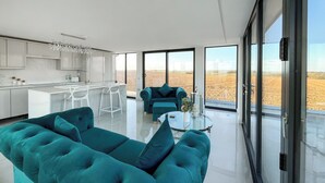 Ein modernes Interieur mit einem petrolfarbenen Sofa, einer Küche mit weißen Schränken und großen Fenstern mit Blick auf ein Feld.