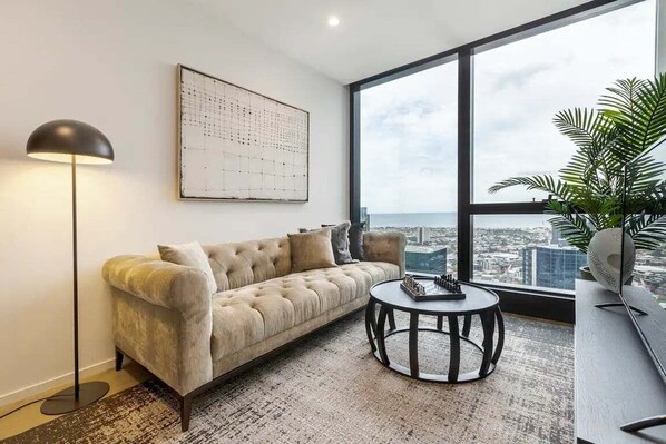 Una sala de estar moderna con un sofá, una mesita redonda, una lámpara de pie, un ventanal con vistas a la ciudad y una planta en maceta.