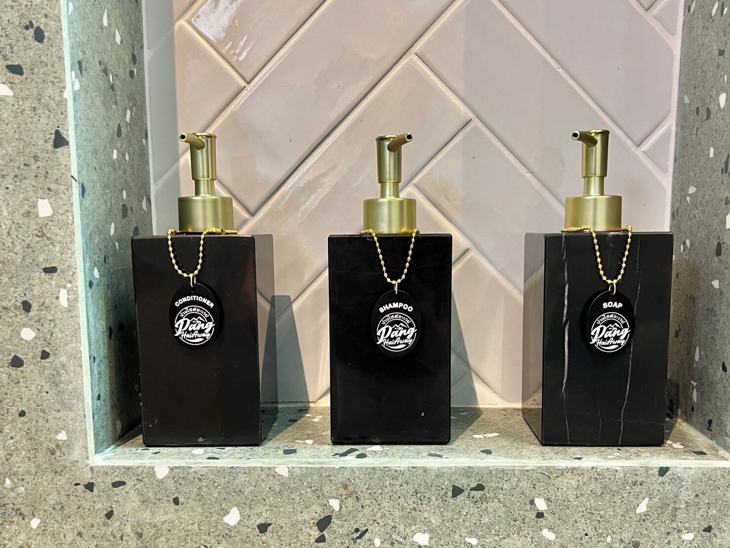 Drei schwarze Flaschen mit goldfarbenen Verschlusskappen und Ketten, jede mit einem anderen Produktnamen beschriftet, präsentiert auf einer gefliesten Ablagefläche.