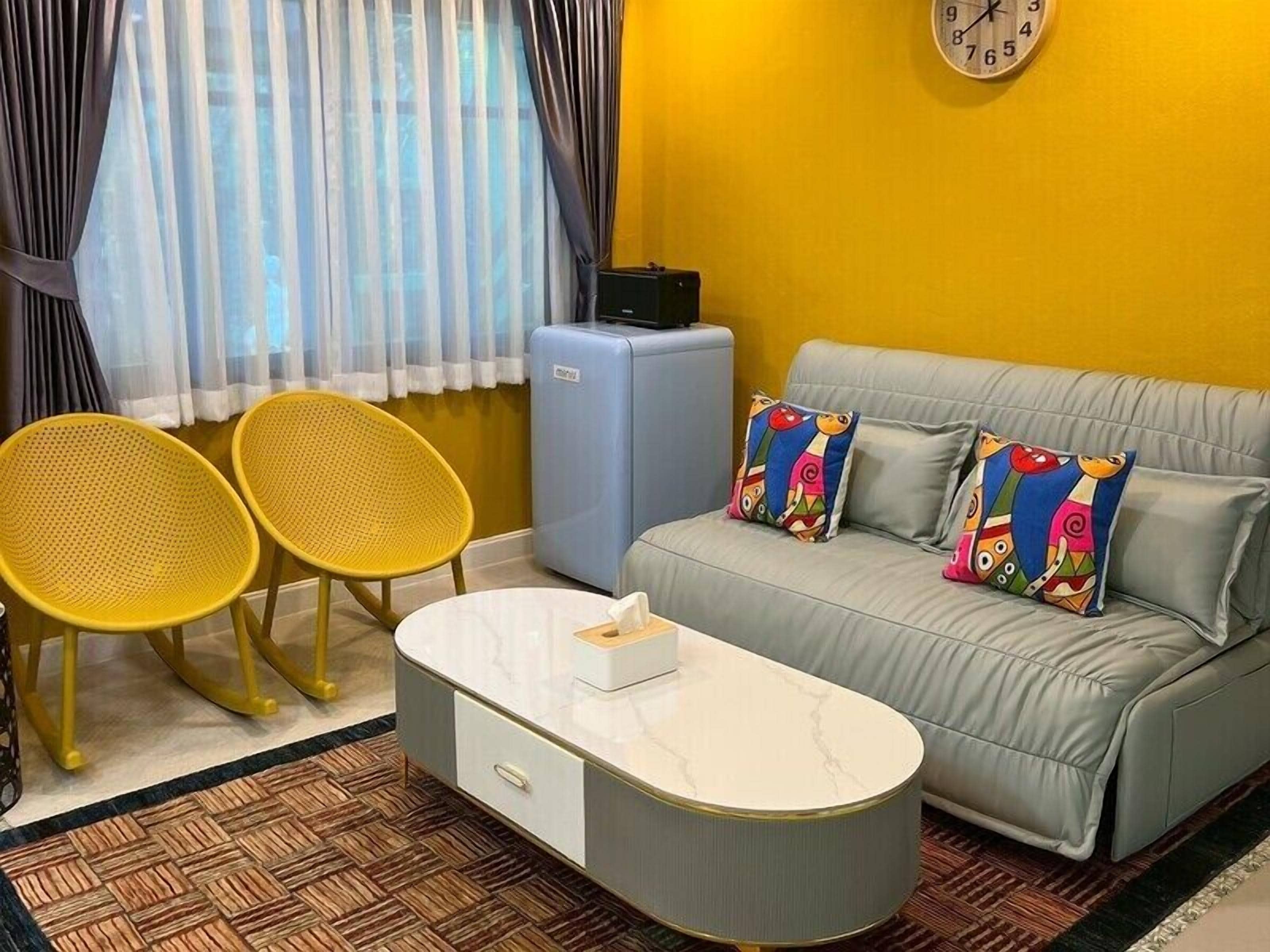 A room with a sofa, two yellow chairs, a small table, a clock on the wall, and a suitcase.