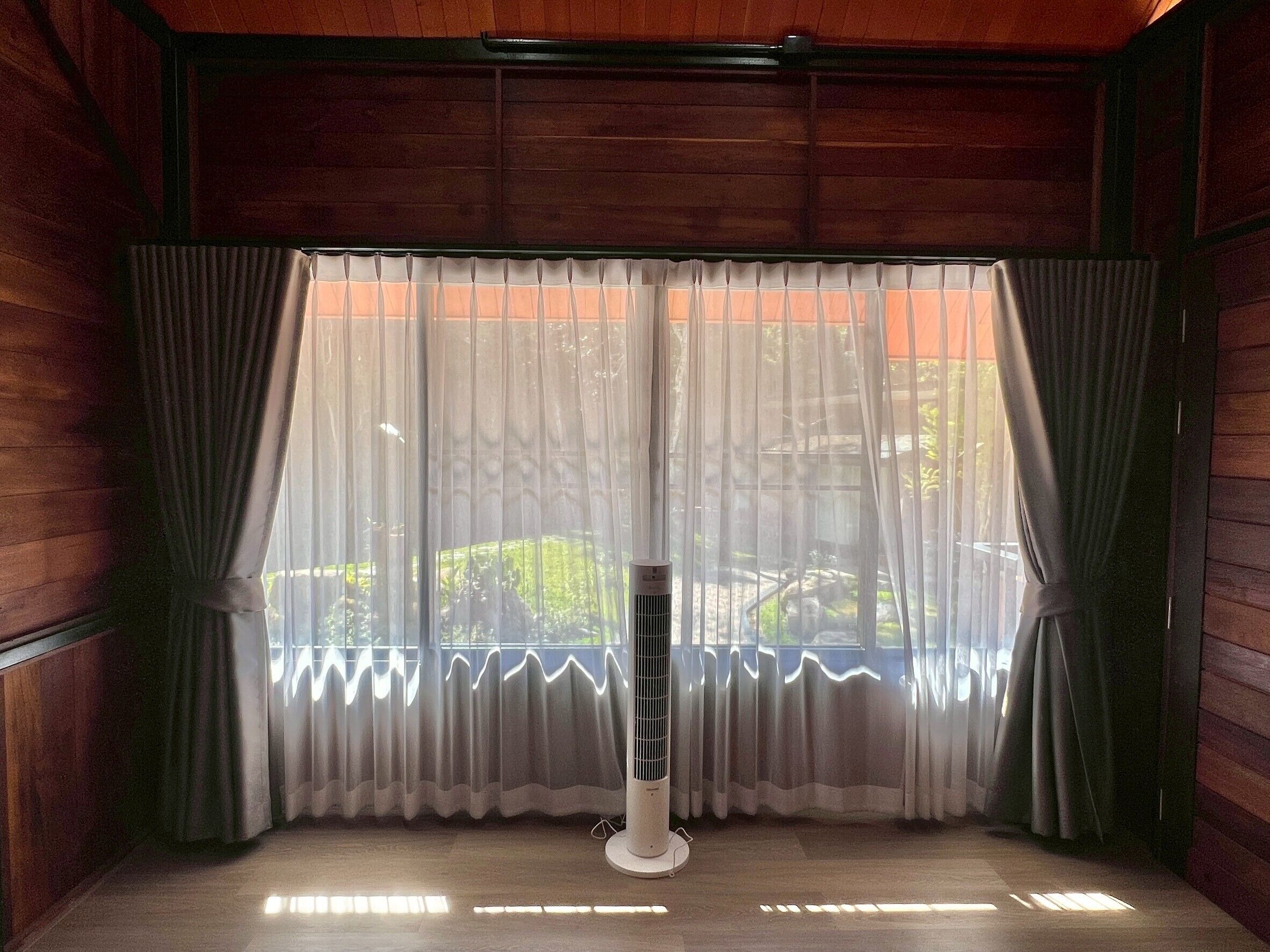 Una habitación con paredes de madera, un ventanal con cortinas traslúcidas y un ventilador de pie.