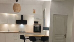 A modern kitchen with white cabinets, a black range hood, and a microwave. There is a dining area with two black chairs and a table. A gray sofa with patterned cushions is in the foreground. - House 5 pers air-conditioned near Avignon/St Remy de Provence air-conditioned parking (Châteaurenard)