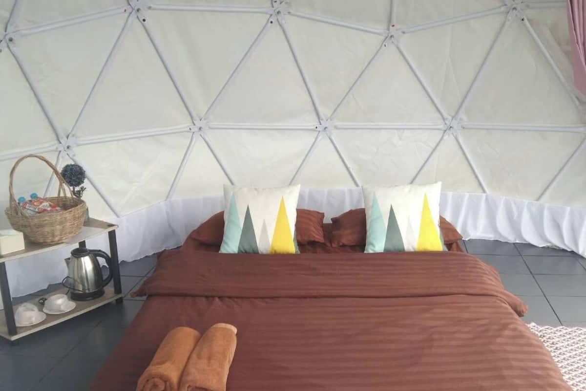 Una habitación tipo tienda de campaña con cama, almohadas, una toalla enrollada, un estante con una cesta y un hervidor.